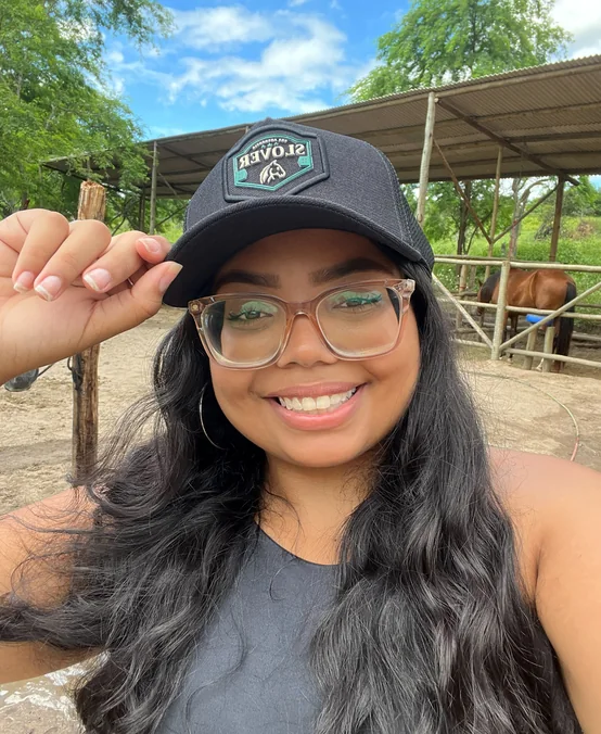Camile Sampaio de boné e óculos em um haras, com um cavalo ao fundo