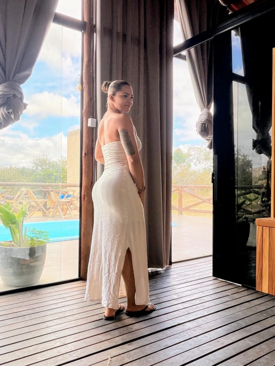 Rutinha, anfitriã do Chalé Vale do Sonho, de vestido branco junto às portas de vidro do chalé, com a piscina ao fundo